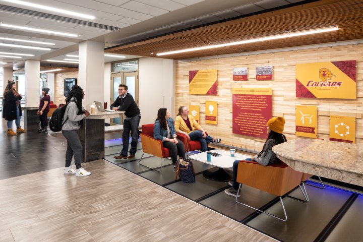 Students and staff gather in a modern student center. Some are seated in a lounge area, talking and relaxing, while others stand at a high table. The walls display informational panels about the university’s history, mission, and values, featuring the University of Minnesota Morris logo.
